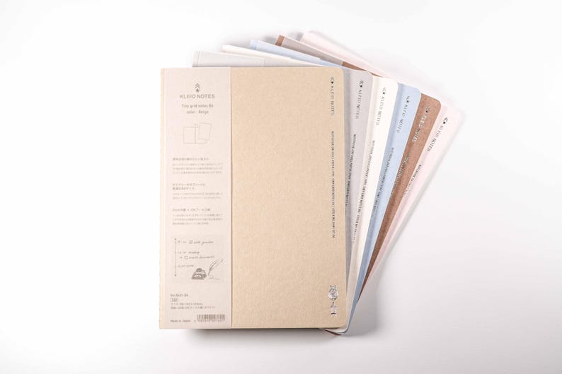K&ouml;nnte beinhalten: Eine Auswahl an Notizb&uuml;chern mit verschiedenfarbigen Einb&auml;nden, darunter Beige, Wei&szlig;, Blau und Braun, f&auml;cherf&ouml;rmig auf wei&szlig;em Hintergrund angeordnet. Das vordere Notizbuch hat ein Etikett mit Text und Illustrationen. Geeignet zum Schreiben oder Skizzieren.
