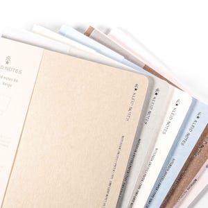 K&ouml;nnte beinhalten: Eine Auswahl an Kleid Notes Notizb&uuml;chern in verschiedenen Farben, darunter Beige, Wei&szlig; und Blau. Die Notizb&uuml;cher haben abgerundete Ecken und den Text "KLEID NOTES" auf der Seite. Auf wei&szlig;em Hintergrund.