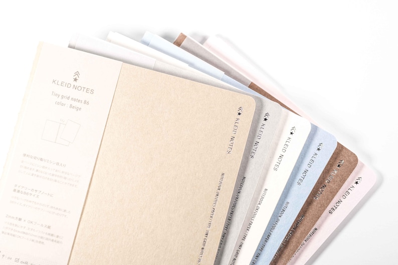 K&ouml;nnte beinhalten: Eine Auswahl an Kleid Notes Notizb&uuml;chern in verschiedenen Farben, darunter Beige, Wei&szlig; und Blau. Die Notizb&uuml;cher haben abgerundete Ecken und den Text "KLEID NOTES" auf der Seite. Auf wei&szlig;em Hintergrund.