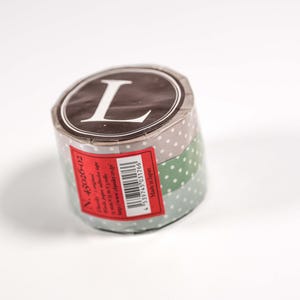 May include: A roll of decorative tape with a brown top featuring a white "L" in a white oval. The tape has layers of gray and green with white polka dots. A red label with text and a barcode is visible.
