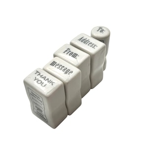 May include: A set of white ceramic stamps with black text. The stamps are stacked and include the words "THANK YOU", "message", "From:", "Address:", and "To:" on their surfaces. The stamps are arranged in a diagonal line.