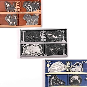 May include: Three sets of decorative paper clips in clear plastic boxes. The top box contains dog-shaped clips, the middle box has dinosaur clips, and the bottom box features space-themed clips. Each box is labeled "16 pcs" and "clips".