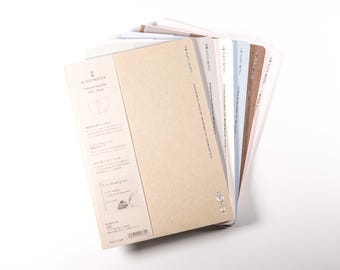 May include: A stack of notebooks with various colored covers, including beige, white, and blue. The notebooks have a simple design and the text "KLEID NOTES" is visible on the cover. The notebooks are fanned out, showing the different colors.