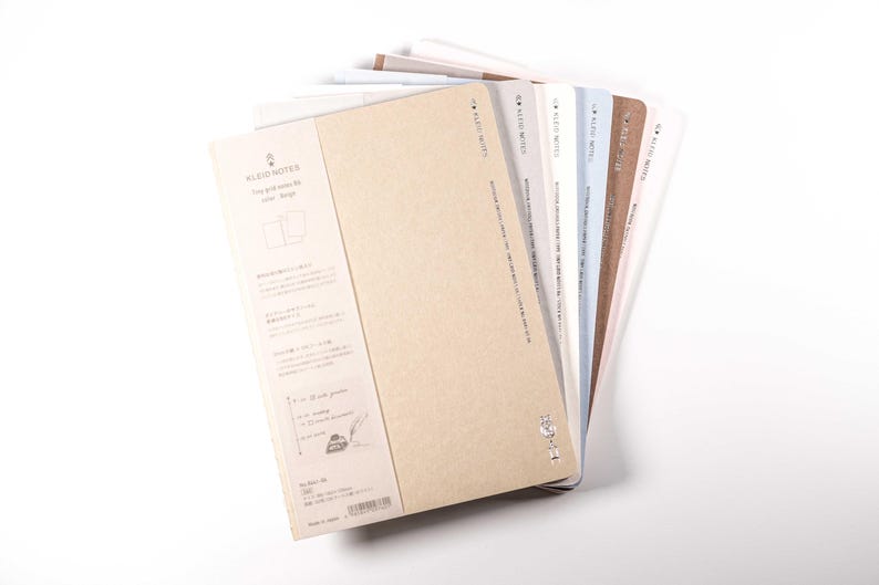 K&ouml;nnte beinhalten: Ein Stapel Notizb&uuml;cher mit verschiedenfarbigen Einb&auml;nden, darunter Beige, Wei&szlig; und Blau. Die Notizb&uuml;cher haben ein schlichtes Design und der Text "KLEID NOTES" ist auf dem Einband zu sehen. Die Notizb&uuml;cher sind aufgef&auml;chert, um die verschiedenen Farben zu zeigen.