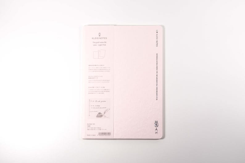 K&ouml;nnte beinhalten: Ein hellrosa Notizbuch mit dem Text "KLEID NOTES" und japanischem Text. Das Notizbuch hat einen weichen Einband und ein schlichtes Design. Das Notizbuch befindet sich auf wei&szlig;em Hintergrund.