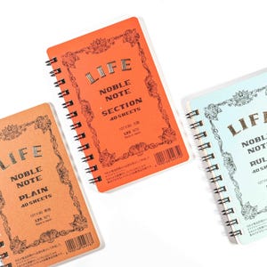 May include: Three spiral-bound notebooks in orange, light orange, and light blue. Each notebook has the word "LIFE" at the top, with "NOBLE NOTE" and "40 SHEETS" below. The orange notebooks say "PLAIN" and "SECTION". The light blue notebook says "RULED".