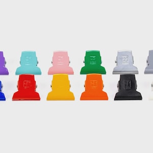 Puede incluir: Una colección de diez clips de encuadernación metálicos de colores, en varios colores, incluyendo morado, azul, rojo, amarillo, naranja, verde, plateado, negro y blanco. Los clips están dispuestos en dos filas, mostrando la parte superior y la base.