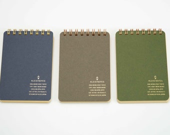 May include: Three small, spiral bound notebooks with gold foil lettering. The notebooks are navy blue, brown, and green. The text on the notebooks reads "KLEID NOTES" and includes details about the paper and design.