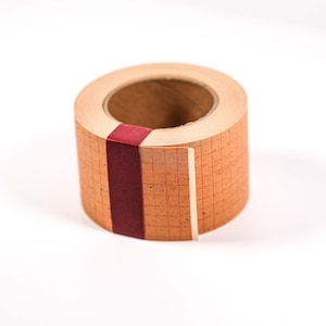 May include: A roll of decorative tape with a grid pattern in shades of tan and brown. A vertical stripe of deep red tape accents the side. The tape is on a white background.