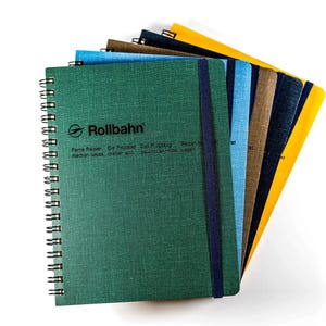 May include: A selection of spiral-bound notebooks in shades of green, blue, and yellow. Each notebook displays the "Rollbahn" logo and text. The notebooks are secured with an elastic band.
