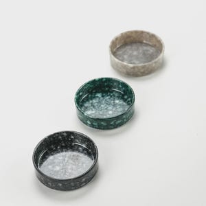 May include: Three small, round ceramic bowls in a row. The bowls are in shades of gray, green, and black, each with a speckled pattern. The bowls are on a white surface.