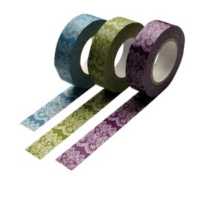 May include: Three rolls of decorative washi tape in blue, green, and purple. Each roll features a white floral lace pattern. The tape is unrolled slightly, showing the pattern. The background is white.