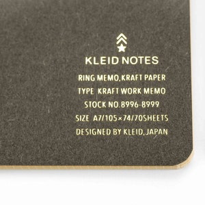 以下が含まれることがあります： 金色文字で「KLEID NOTES」と書かれた茶色の紙製ノート。リングメモで、クラフト紙製。KLEID, JAPANのデザイン。在庫番号は8996-8999、サイズはA7/105&times;74/70枚です。