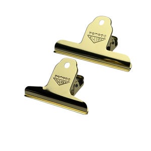 May include: Two gold-coloured metal binder clips with a shiny finish. Each clip features a triangular tab at the top with a circular hole and the embossed text "penco" above the word "CLAMP" inside a triangular border design on the front surface.