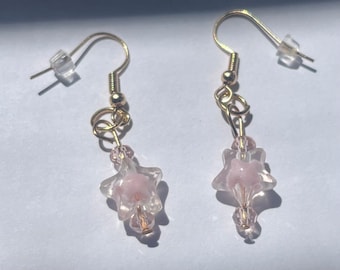 May include: A pair of gold dangle earrings with pink star-shaped glass beads. The beads are clear with a pink swirl inside.