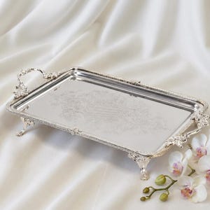 May include: A rectangular silver-toned serving tray with ornate, raised floral handles and four decorative feet. The center of the tray features a subtle, etched floral pattern, and the edges are finished with a detailed, beaded border design.