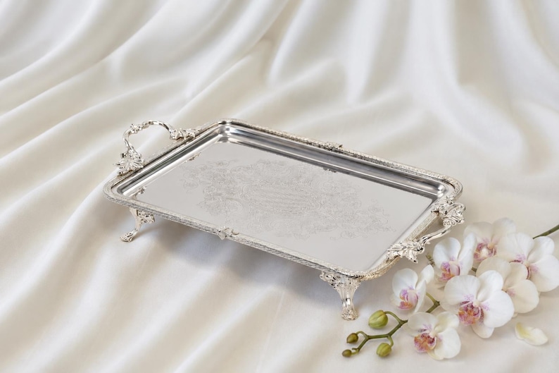 May include: A rectangular silver-toned serving tray with ornate, raised floral handles and four decorative feet. The center of the tray features a subtle, etched floral pattern, and the edges are finished with a detailed, beaded border design.