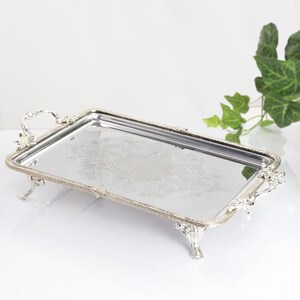 May include: A rectangular silver-toned serving tray with a mirrored base featuring an intricate etched floral pattern. The tray has ornate handles on both ends and stands on four decorative feet with a beaded edge along the entire perimeter.