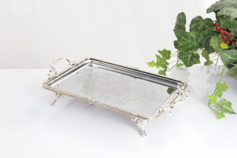 May include: A rectangular silver-toned serving tray with a mirrored base featuring an intricate etched floral pattern. The tray has ornate handles on both ends and stands on four decorative feet with a beaded edge along the entire perimeter.