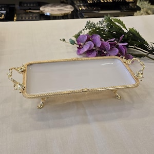 May include: A rectangular serving tray with a white interior and a gold-colored, ornate frame. The tray has decorative handles and rests on four small, curved feet. The frame features a detailed, textured design. Purple flowers and greenery are in the background.