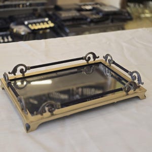 Pode incluir: Uma bandeja retangular com espelho, apresentando uma moldura de metal dourado com quatro detalhes de canto ornamentados em forma de voluta e barras horizontais pretas finas que conectam os postes decorativos laterais, criando uma borda.