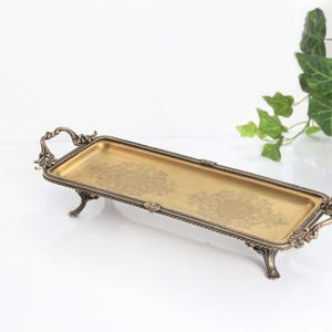 May include: A rectangular gold-toned metal tray with an ornate, dark bronze-colored border and matching handles on each end. The tray features four decorative legs and a subtle, etched floral pattern on the flat, metallic gold surface.