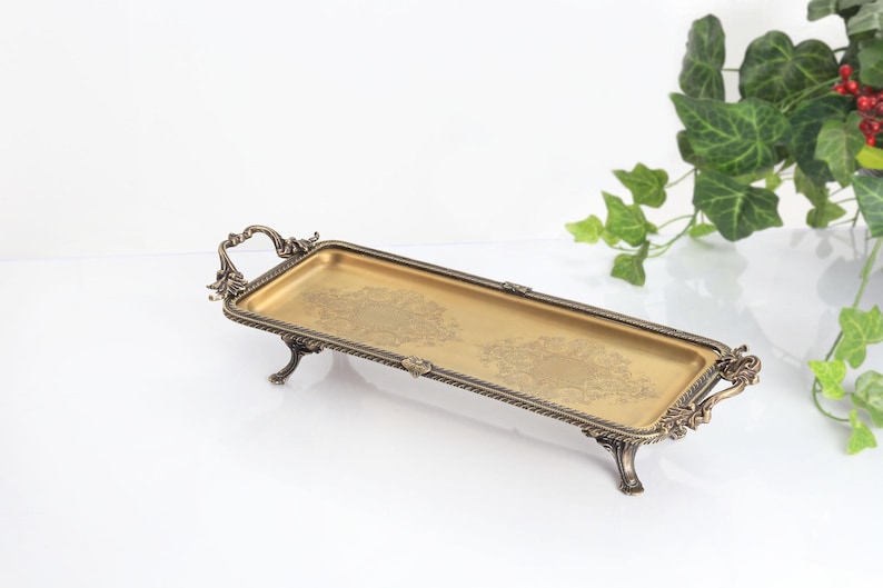 May include: A rectangular gold-toned metal tray with an ornate, dark bronze-colored border and matching handles on each end. The tray features four decorative legs and a subtle, etched floral pattern on the flat, metallic gold surface.