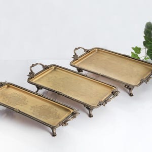 May include: Three rectangular gold-toned metal trays with ornate, dark bronze-colored handles and feet. Each tray features a flat base with a subtle, intricate floral pattern etched into the center, elevated by four decorative legs and side handles.