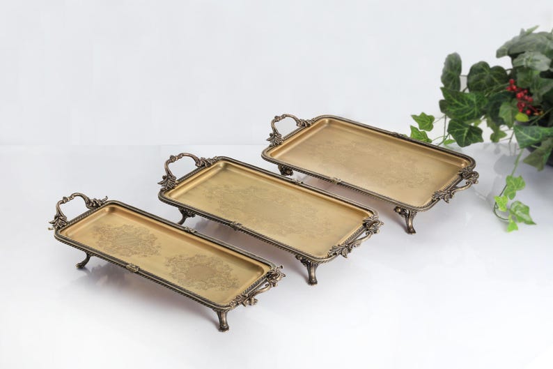 May include: Three rectangular gold-toned metal trays with ornate, dark bronze-colored handles and feet. Each tray features a flat base with a subtle, intricate floral pattern etched into the center, elevated by four decorative legs and side handles.