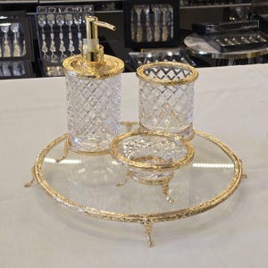 May include: A luxurious bathroom set featuring a soap dispenser, tumbler, and soap dish, all in clear glass with a diamond pattern and gold-colored accents. The set sits on a round glass tray with a gold-colored ornate frame and feet. A sophisticated home decor item.