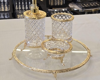 May include: A luxurious bathroom set featuring a soap dispenser, tumbler, and soap dish, all in clear glass with a diamond pattern and gold-colored accents. The set sits on a round glass tray with a gold-colored ornate frame and feet. A sophisticated home decor item.