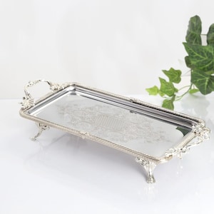 May include: A rectangular silver-toned serving tray with a reflective mirrored surface, featuring ornate decorative handles and four raised feet. The edges of the tray are detailed with a beaded texture, and the center displays an intricate etched pattern.