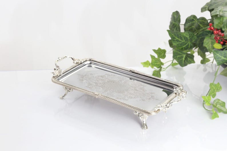 May include: A rectangular silver-toned serving tray with a reflective mirrored surface, featuring ornate decorative handles and four raised feet. The edges of the tray are detailed with a beaded texture, and the center displays an intricate etched pattern.