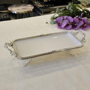 Op de afbeelding: Een rechthoekige serveerschaal met een witte binnenkant en een sierlijk zilverkleurig metalen frame. De schaal heeft decoratieve handgrepen en staat op kleine voetjes. Het ontwerp is elegant en geschikt voor het serveren van eten of het presenteren van items.
