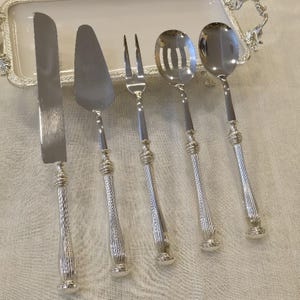 Könnte beinhalten: Ein Set aus fünf silbernen Servierutensilien, darunter ein Messer, ein Tortenheber, eine Gabel, ein Schaumlöffel und ein Löffel. Jedes Teil hat einen dekorativen Griff mit geripptem Design und aufwendigen Details. Das Set liegt auf einem weißen Serviertablett.