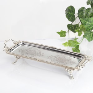 May include: A rectangular silver-toned metal serving tray featuring ornate, raised decorative handles and four detailed feet. The flat surface of the tray displays two symmetrical, intricate etched floral patterns centered on the polished metal base.