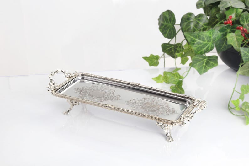 May include: A rectangular silver-toned metal serving tray featuring ornate, raised decorative handles and four detailed feet. The flat surface of the tray displays two symmetrical, intricate etched floral patterns centered on the polished metal base.