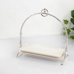 Può includere: Un supporto da portata in metallo color argento caratterizzato da un'alta maniglia ad arco con un anello decorativo sulla sommità. Il supporto sostiene un vassoio rettangolare in ceramica bianca con bordi leggermente rialzati, fissato da due montanti verticali.