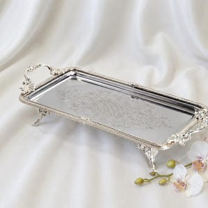 May include: A rectangular silver-toned metal tray with a reflective surface, featuring ornate handles and four decorative feet. The edges of the tray are detailed with a beaded texture, and the center displays an intricate, etched floral pattern.