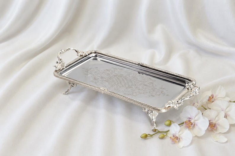 May include: A rectangular silver-toned metal tray with a reflective surface, featuring ornate handles and four decorative feet. The edges of the tray are detailed with a beaded texture, and the center displays an intricate, etched floral pattern.