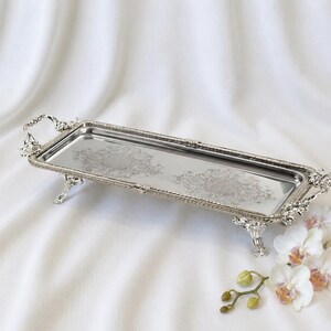 May include: A rectangular silver-toned serving tray with ornate handles and four decorative feet. The tray features a polished, reflective surface with intricate engraved floral patterns along the center and a textured, beaded border edge.