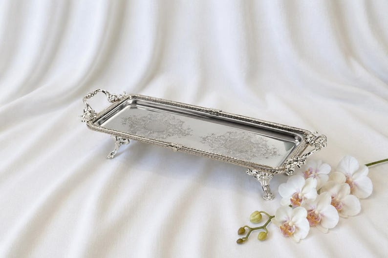 May include: A rectangular silver-toned serving tray with ornate handles and four decorative feet. The tray features a polished, reflective surface with intricate engraved floral patterns along the center and a textured, beaded border edge.