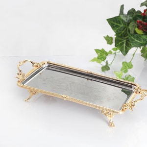 May include: A rectangular serving tray featuring a reflective mirrored surface and an ornate gold-toned metal frame. The tray is supported by four decorative legs and includes two matching gold-toned handles on the short ends of the frame.