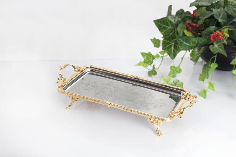 May include: A rectangular serving tray featuring a reflective mirrored surface and an ornate gold-toned metal frame. The tray is supported by four decorative legs and includes two matching gold-toned handles on the short ends of the frame.