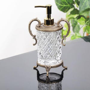 May include: A clear glass soap dispenser with a diamond-cut pattern. It has a gold-colored pump and ornate metal accents. The dispenser is supported by a decorative base with three legs and two handles. A stylish bathroom accessory.