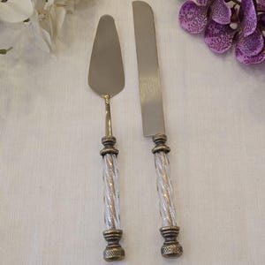 May include: A cake serving set consisting of a flat triangular server and a long straight knife. Both utensils feature metallic silver blades and clear, spiraled glass handles with antique brass-toned metal accents at the top and bottom.