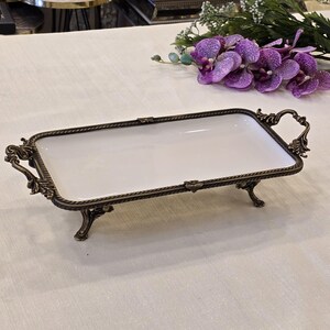 May include: A rectangular serving tray with a white interior and an ornate bronze-coloured metal frame. The tray has decorative handles and stands on four small feet. The frame features a rope-like border and floral accents.