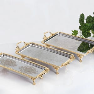 May include: A set of three rectangular mirrored trays with ornate gold-toned metal frames, decorative handles, and four small feet. Each tray features a reflective surface with a central etched floral pattern and a polished metallic border.
