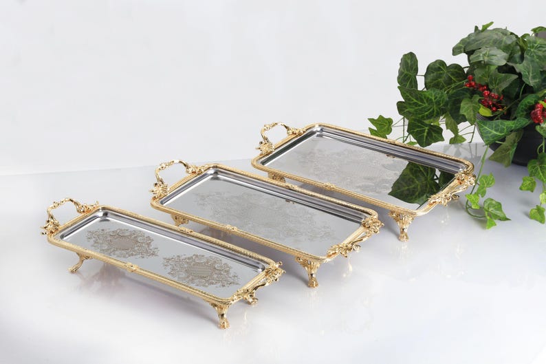 May include: A set of three rectangular mirrored trays with ornate gold-toned metal frames, decorative handles, and four small feet. Each tray features a reflective surface with a central etched floral pattern and a polished metallic border.