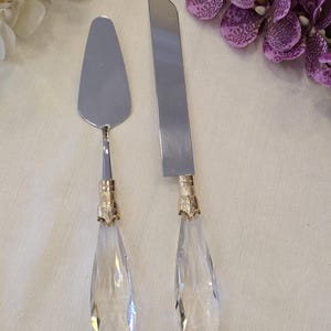 Può includere: Un set di coltello e paletta per torte argentati. I manici sono in cristallo trasparente a forma di goccia con dettagli dorati. Il server ha una lama larga e piatta e il coltello ha una lama lunga e dritta. Il set è progettato per servire la torta.